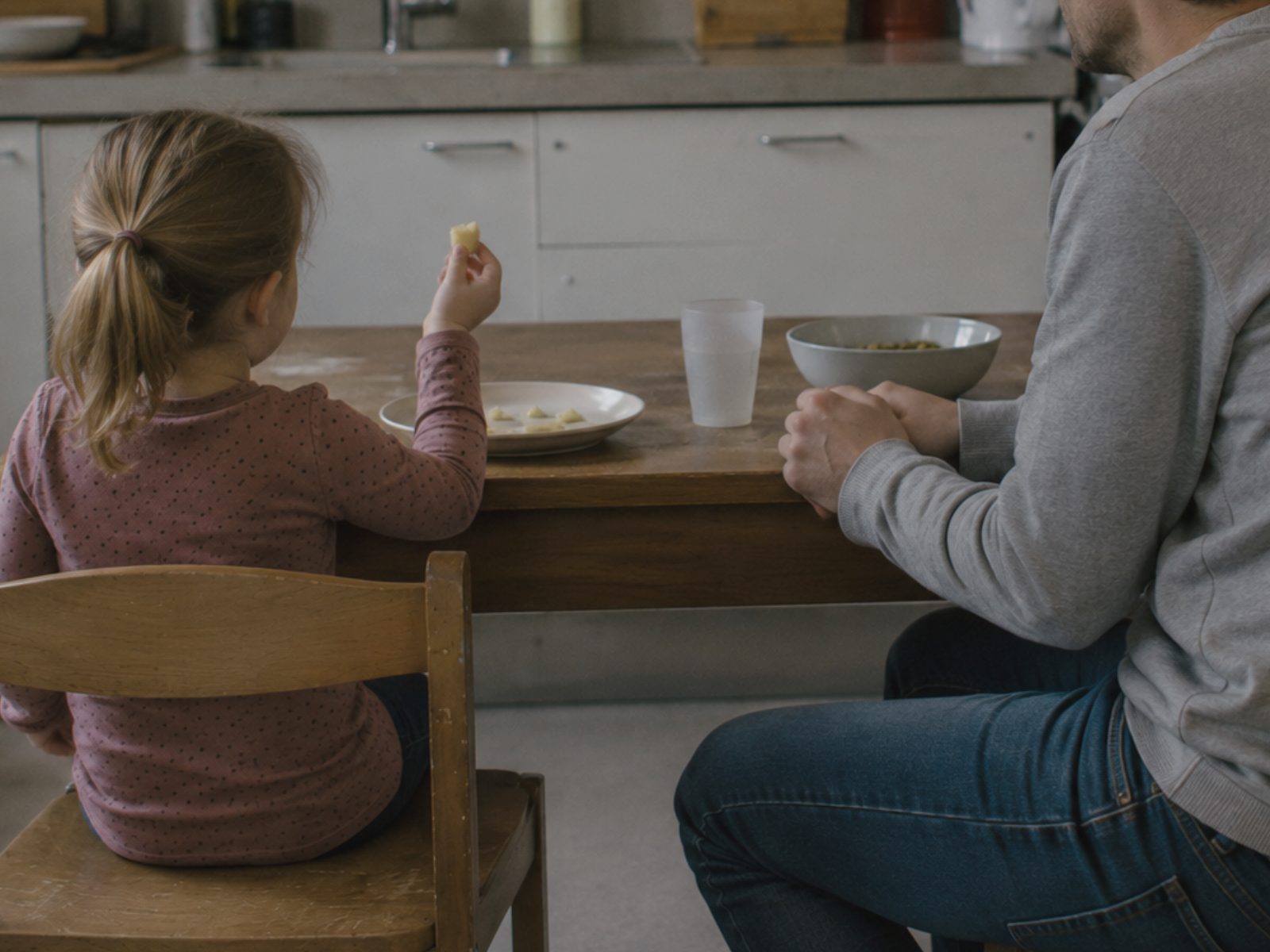 Een meisje houdt een stukje eten stil in haar hand en kijkt ernaar; naast haar zitten de handen van haar vader gevouwen op tafel