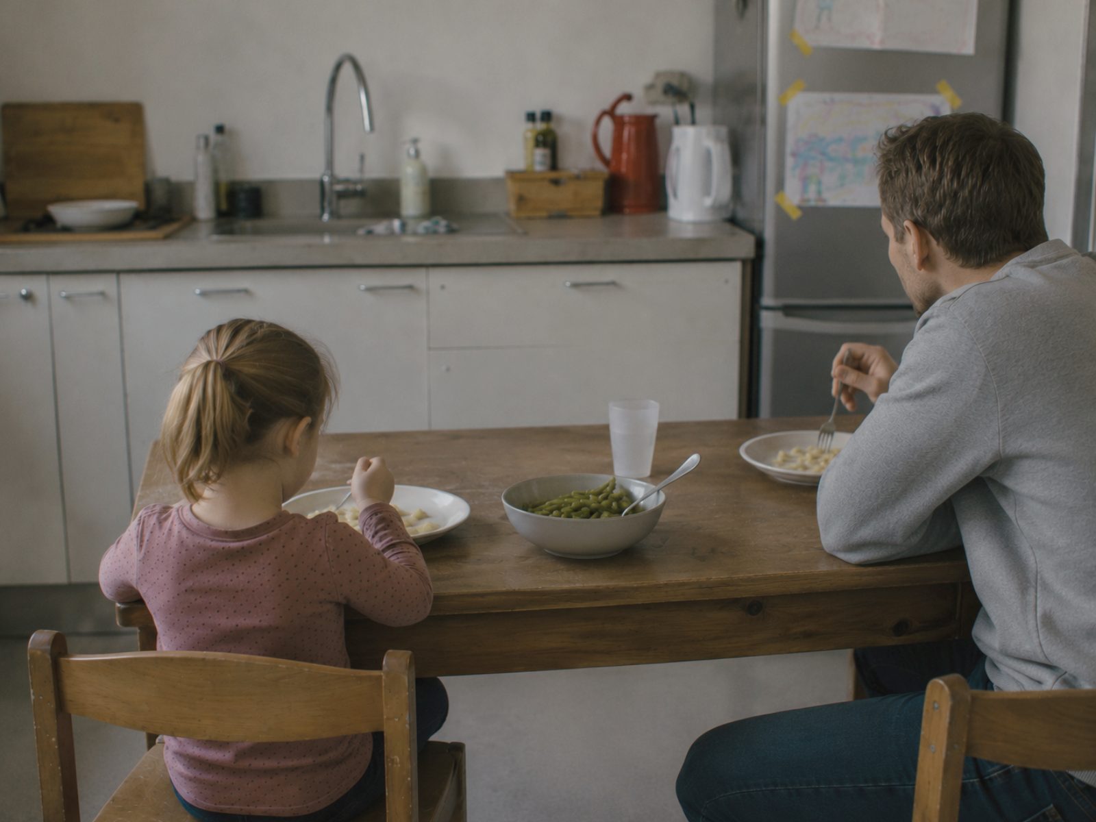 Een meisje eet pasta van haar eigen bord terwijl de gezamenlijke schaal met groente onaangeroerd naast haar staat; haar vader eet zijn eigen eten en kijkt niet naar haar bord